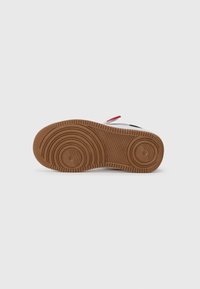 Semelle de chaussure avec des motifs circulaires texturés en caoutchouc marron. Présente des accents rouges et noirs sur un dessus blanc. Design plat et profil bas.