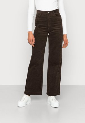 Mujer con pantalones de pana anchos de pierna en tono marrón oscuro con bolsillos delanteros, combinados con zapatillas blancas de plataforma y un top blanco ajustado de canalé.