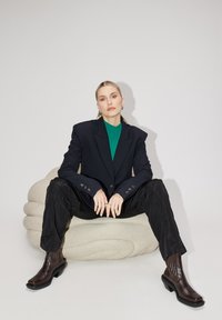 Blazer noir ajusté avec des épaules larges, top en tricot vert, pantalon noir ample et bottes à talons marron sur un coussin beige texturé.