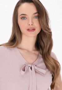 Top rosa chiaro con scollo a V, caratterizzato da un dettaglio a fiocco legato al colletto. Tessuto morbido, dal minimo lucido e maniche corte.
