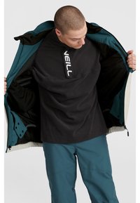Chaqueta de abrigo para hombre, con un interior en verde azulado y detalles en blanco, cierre con cremallera, forro aislante, llevada abierta sobre un sudadera negra y pantalones en verde azulado.