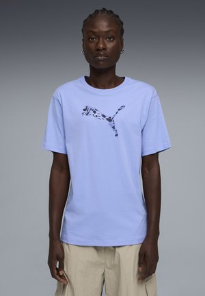 Person trägt ein hellviolettes Puma-T-Shirt mit einem gefleckten Puma-Logo und beige Cargohosen, steht vor einem einfarbigen grauen Hintergrund.