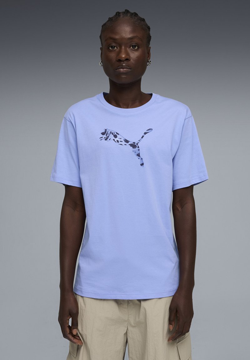 Person iført en lys lilla Puma t-shirt med en pletvis Puma-logo og beige cargobukser, stående mod en ensfarvet grå baggrund.