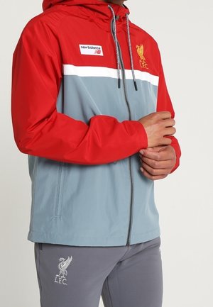 Osoba nosząca czerwoną i szarą kurtkę na suwak z logo Liverpool FC i New Balance, dłonie splecione z przodu, ubrana w szare spodnie Liverpool FC.