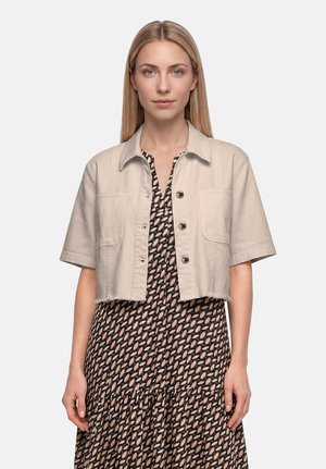 Femme aux cheveux blonds raides portant une veste beige courte boutonnée par-dessus une robe à motifs, debout devant un fond uni.