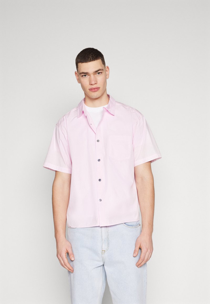 Camisa de manga corta abotonada en rosa pastel, con un bolsillo en el pecho y botones negros. Hecha de una tela ligera con una textura suave.