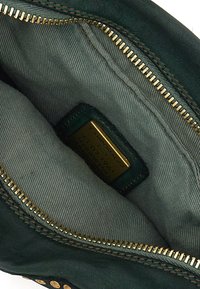Interno di una borsa in pelle verde con foderatura in tessuto grigio chiaro. Cerniera dorata e un cartellino dorato con i dettagli del marchio all'interno.