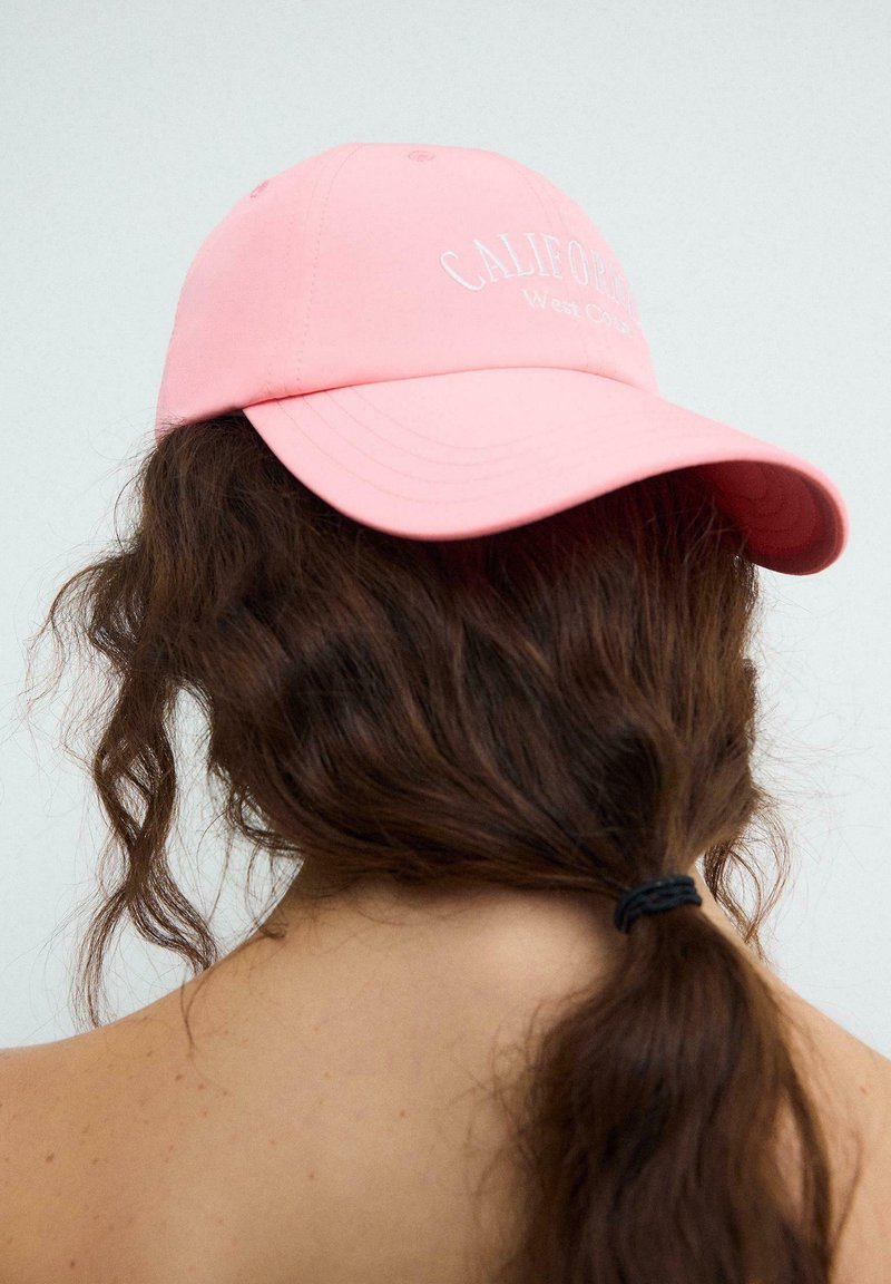 Persona con capelli lunghi e scuri raccolti che indossa un berretto rosa con la scritta "California West Coast", vista da dietro su uno sfondo chiaro.