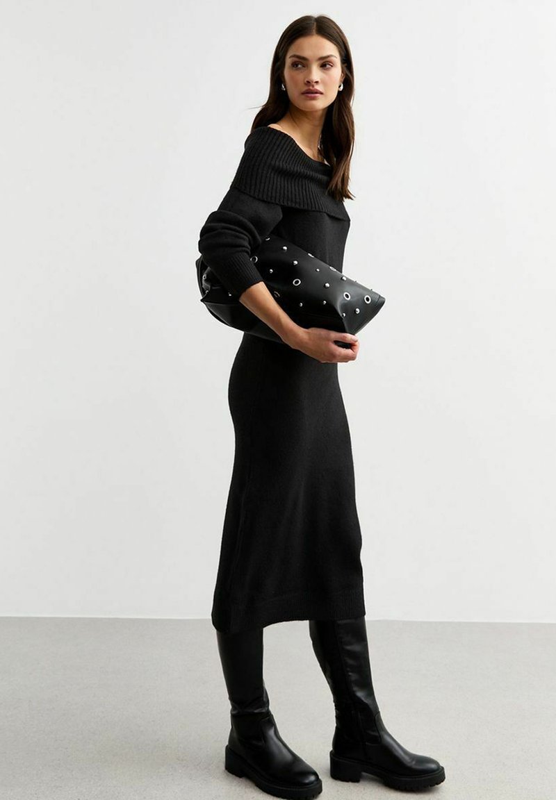 Abito nero lavorato a maglia con colletto scollato, abbinato a stivali neri al ginocchio. Tiene in mano una pochette nera con dettagli in metallo.