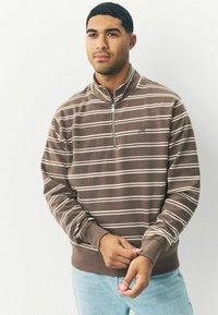 Brunstribet zip-up sweatshirt med hvide striber, ribbede manchetter og bund. Har et diskret logo på brystet. Bæres med lyseblå jeans.