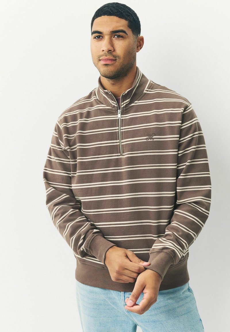 Brunstribet zip-up sweatshirt med hvide striber, ribbede manchetter og bund. Har et diskret logo på brystet. Bæres med lyseblå jeans.