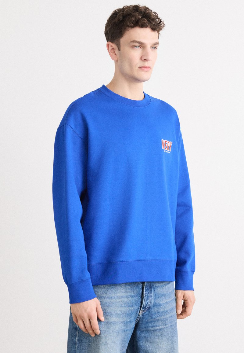 Sweatshirt azul com punhos e barra canelados, apresentando um pequeno gráfico em texto laranja no lado esquerdo do peito. Material macio e corte descontraído.
