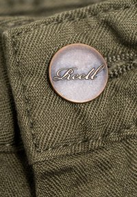 Bouton métallique rond avec le mot "Reell" en cursive, fixé à un tissu vert texturé avec des coutures visibles.