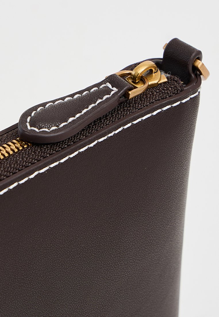Primo piano di una borsa in pelle marrone scuro che mostra una zip dorata e un tirante con cuciture bianche a contrasto lungo i bordi.