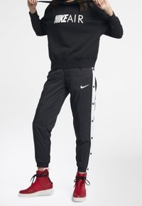 Svart sweatshirt med "NIKE AIR"-logotyp, kombinerad med svarta byxor med vita sidoknappar och röda högklackade sneakers.