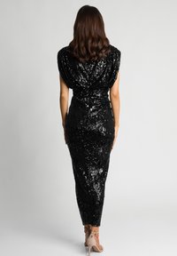 Kobieta z falowanymi brązowymi włosami ma na sobie czarną sukienkę midi z cekinami oraz beżowe sandały na wysokim obcasie, stoi na tle gładkiego białego tła.