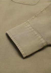 Camicia beige chiaro con una trama liscia, dotata di colletto con bottoni e polsini ben cuciti. La vista ravvicinata mette in evidenza i dettagli del tessuto.