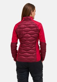 Vrouw met de rug naar de camera in een kastanjebruine en rode gewatteerde jas, met zwarte gewatteerde broek, tegen een effen achtergrond.