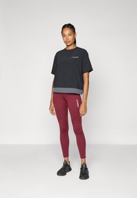 Camiseta deportiva corta negra con dobladillo gris, combinada con leggings burdeos que presentan un logo lateral, y zapatos deportivos negros. Textura de tela suave.