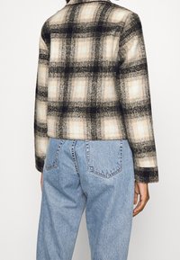 Kurtka w kratę w odcieniach czerni, beżu i szarości. Charakteryzuje się miękką fakturą, kołnierzem i długimi rękawami, noszona na jasnych, dżinsowych spodniach.