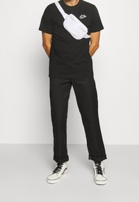 Svart T-shirt med vitt Nike-logga, lösa svarta byxor och vit midjeväska bärs över kroppen. Vita sneakers med svarta snören.