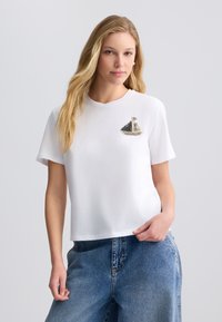 Hvid bomulds t-shirt med et lille dekorativt båddesign i sort, grå og guld palietter på venstre bryst. Korte ærmer.
