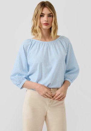 Kobieta o blond włosach do ramion, ubrana w niebiesko-białą bluzkę w paski z rękawami do łokci oraz beżowe spodnie.