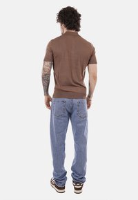 Hombre con cabello rizado y tatuajes viste un polo marrón de manga corta, jeans azul claro y zapatillas Nike marrones, de pie mirando hacia atrás.