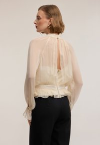 Blusa cremosa chiara in tessuto trasparente, maniche gonfie e vita con volant. Presenta una cerniera posteriore e un dettaglio con bottone sul colletto.