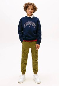 Ragazzo sorridente con capelli ricci che indossa una felpa Tommy blu navy, pantaloni cargo verde oliva e sneakers bianche, in piedi con le mani nelle tasche.