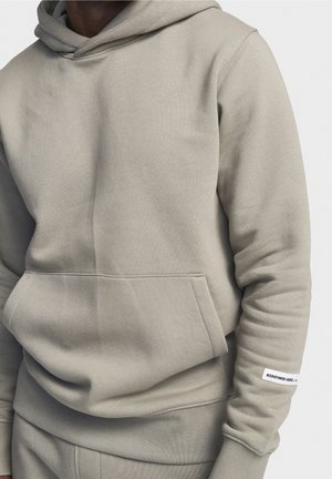 Lys grå hættetrøje lavet af blødt, struktureret stof. Har en frontlomme, snørebånd i hætten og et mærke på ærmet med sort tekst.