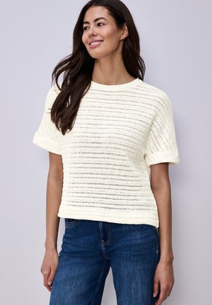 Smilende kvinde med langt mørkt hår iført en hvid kortærmet striktrøje og blå jeans foran en ensartet lys grå baggrund.