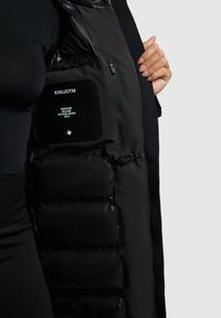 Svart puffväst med en glänsande yta och quiltade sektioner. Innehåller en dragkedjeförsedd ficka med ett varumärkesmärke inuti.