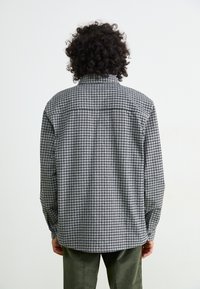 Persoon met krullend haar, gekleed in een zwart-wit geruit overhemd met lange mouwen en olijfgroene broek, met de rug naar de camera tegen een effen achtergrond.