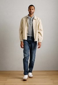 Man wearing beige jacket, gray polo shirt, blue jeans, and white sneakers walking on wooden floor against plain light wall.