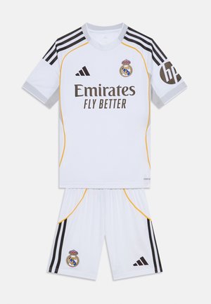 Bela nogometna uniforma s kratkimi rokavi in kratkimi hlačami, z blagovnimi znaki črne in rumene barve, logotipom Adidas ter grbom Real Madrida na majici in hlačah.