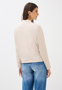 Lichtbeige jas, lichtgewicht materiaal, ontspannen pasvorm, ribgebreide boorden en tailleband, met een eenvoudige naden detail aan de achterkant. Gecombineerd met blauwe jeans.