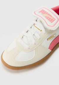 Baskets Puma Palermo avec une tige en synthétique blanc, accents roses et logo doré. Présente un système de laçage et une semelle en gomme texturée.