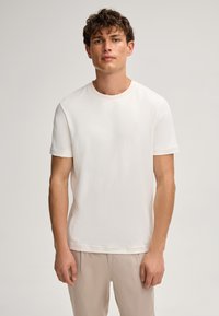 Witte, korte mouwen t-shirt van zachte stof met een ronde halslijn en een subtiele, reliëf logo op de borst. Gecombineerd met licht beige broeken.
