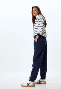 Hvid hoodie med marineblå striber, parret med marineblå sweatpants og hvide sneakers med sorte accenter. Modellen står med hænderne i lommerne.