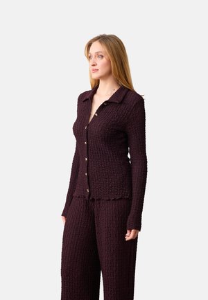 Kobieta ubrana w teksturowaną, ciemnofioletową bluzkę na guziki z długim rękawem i pasujące szerokie spodnie, stojąca na tle jednolitego, jasnego tła.