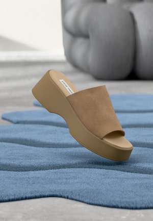 Sandale plateforme beige avec dessus en daim et bout ouvert, posée sur un tapis bleu texturé avec du mobilier capitonné gris en arrière-plan.
