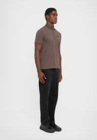 Brun kortermet polo skjorte med krage og brodert logo, kombinert med svarte bukser og svarte joggesko, stående på en hvit bakgrunn.