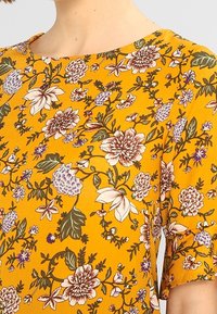 Žlutá látka s podrobným květinovým vzorem v odstínech bílé, fialové, zelené a hnědé na krátkém rukávu.
