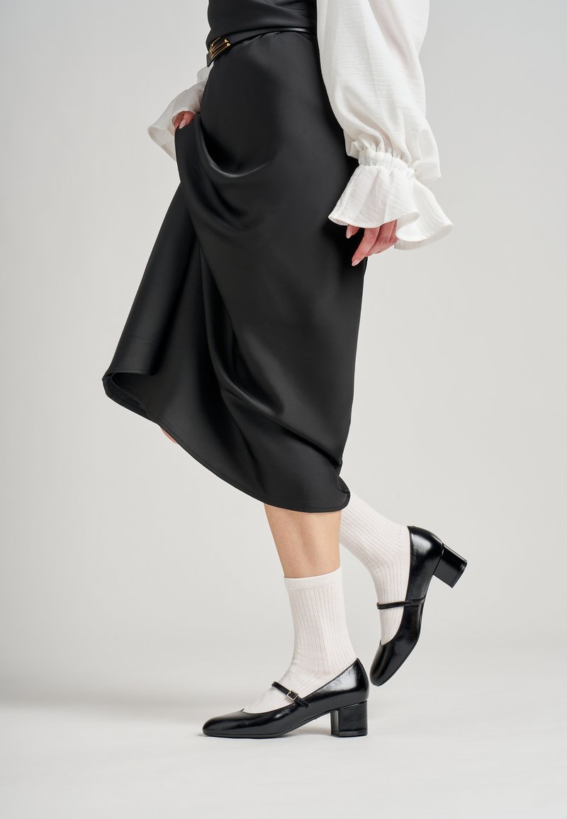 Falda A-line de satén negro con un brillo sutil, combinada con calcetines blancos de canalé y zapatos de charol negro con tacón cuadrado y una correa.