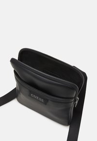 Schwarze Leder-Crossbody-Tasche mit Reißverschluss. Verfügt über eine Vordertasche und einen Textilgurt. Markenname auf der Vorderseite eingeprägt.