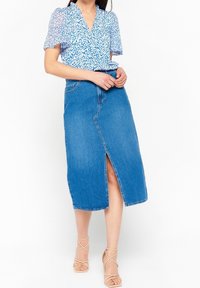 Una donna indossa una blusa blu a fiori e una gonna in denim a lunghezza ginocchio con uno spacco frontale, abbinata a tacchi beige con cinturini.