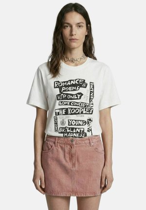 Jeune femme portant un t-shirt blanc à motif avec texte noir et une jupe en denim rose, debout devant un fond blanc uni.