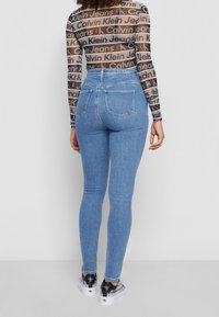 Modré skinny džíny s vysokým pasem a zadními kapsami. Modelka má na sobě průhledný top s dlouhým rukávem, který má opakující se vzor loga.