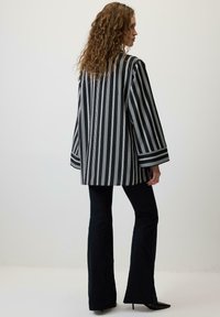 Ženska s kodrastimi lasmi, oblečena v črno-belo veliko jopico z navpičnimi črtami, črne zvonaste hlače in črne koničaste pete, stoji postrani.